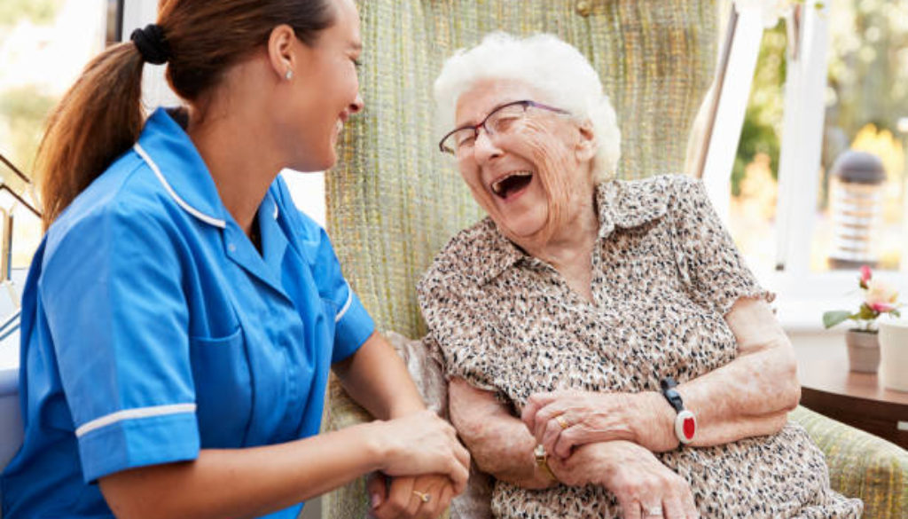 Senior Woman Sitting In Chair And Laughing With Nurse In Retirement Home